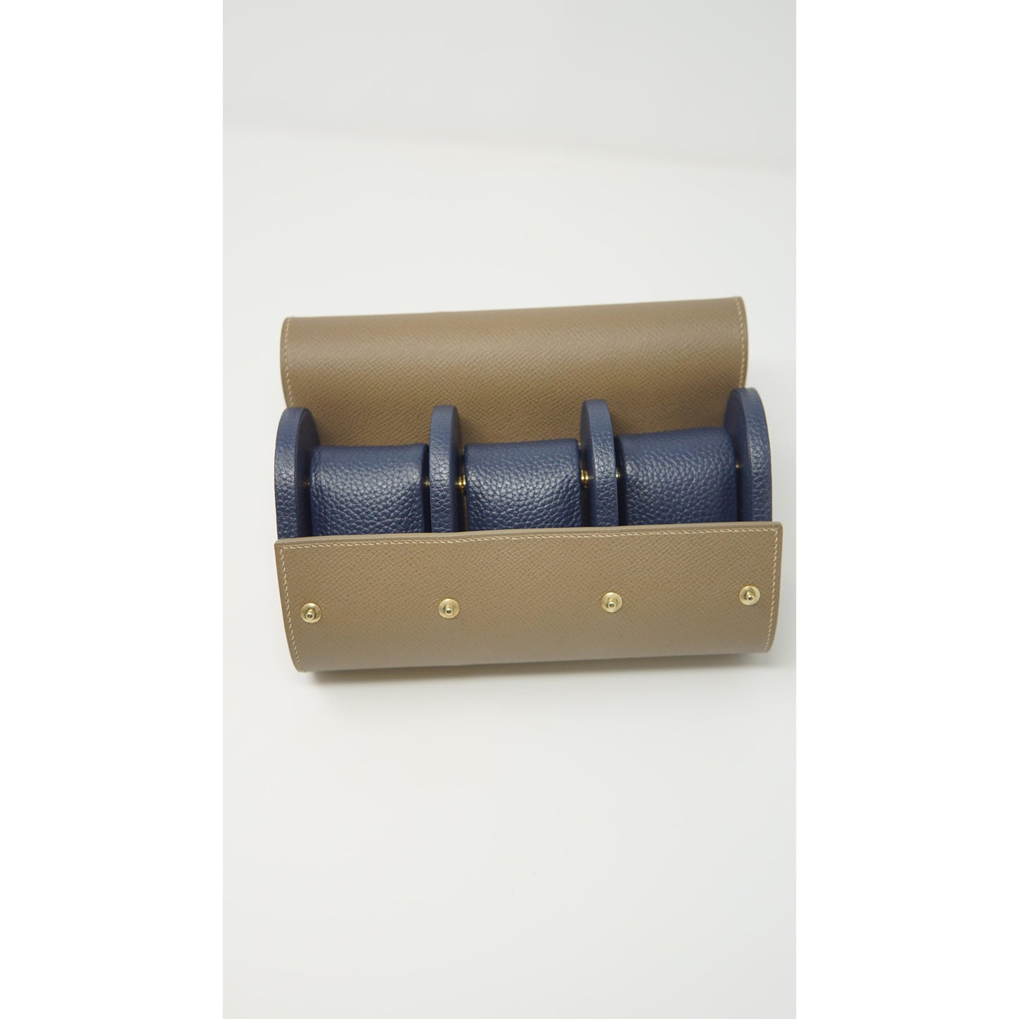 Tan leather watch roll with three navy blue leather watch strap holders and gold snap closures, flat lay.