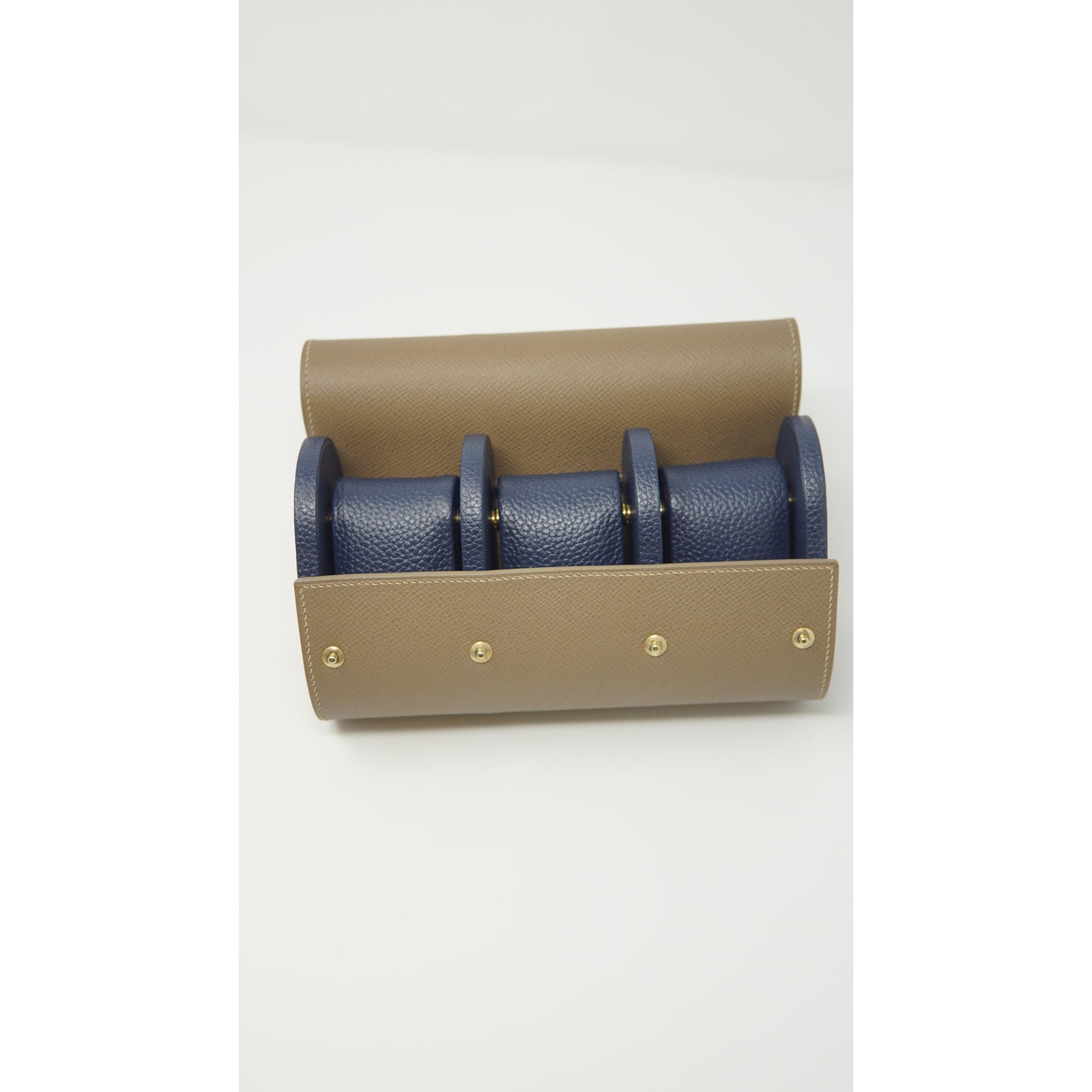 Tan leather watch roll with three navy blue leather watch strap holders and gold snap closures, flat lay.