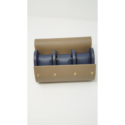 Tan leather watch roll with three navy blue leather watch strap holders and gold snap closures, flat lay.