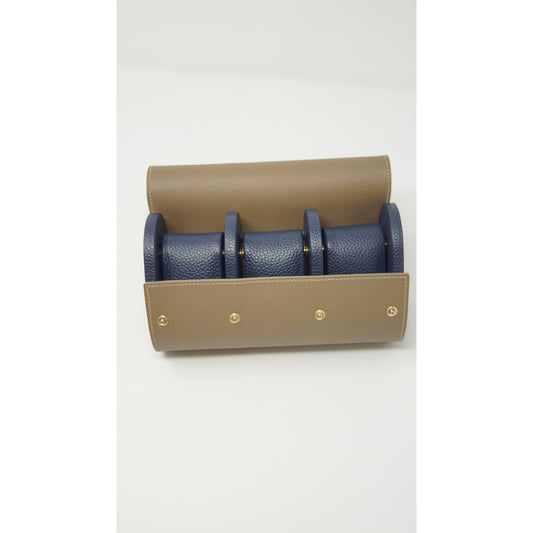 Tan leather watch roll with three navy blue leather watch strap holders and gold snap closures, flat lay.