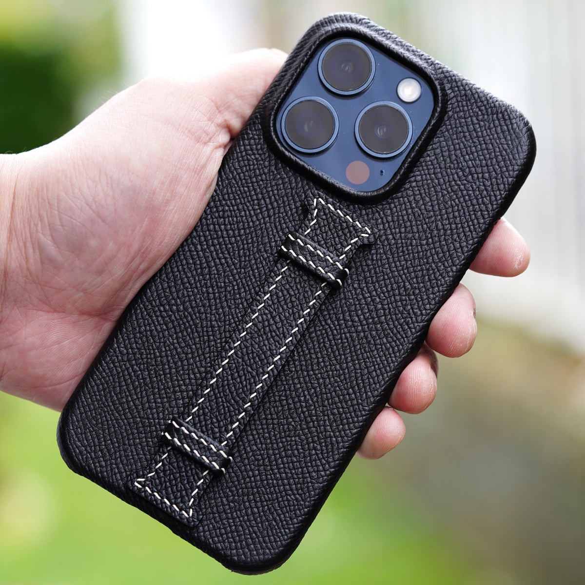 Black Epsom leather iPhone case with white stitched grip, held in hand.
