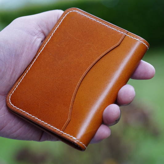 Hand holding a tan leather card holder with white stitching and a curved pocket design.