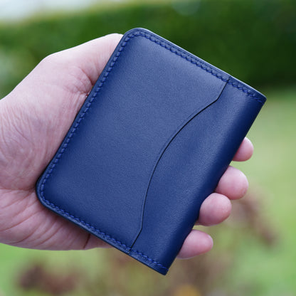 Hand holding a blue leather card holder with matching stitching and a curved front pocket.