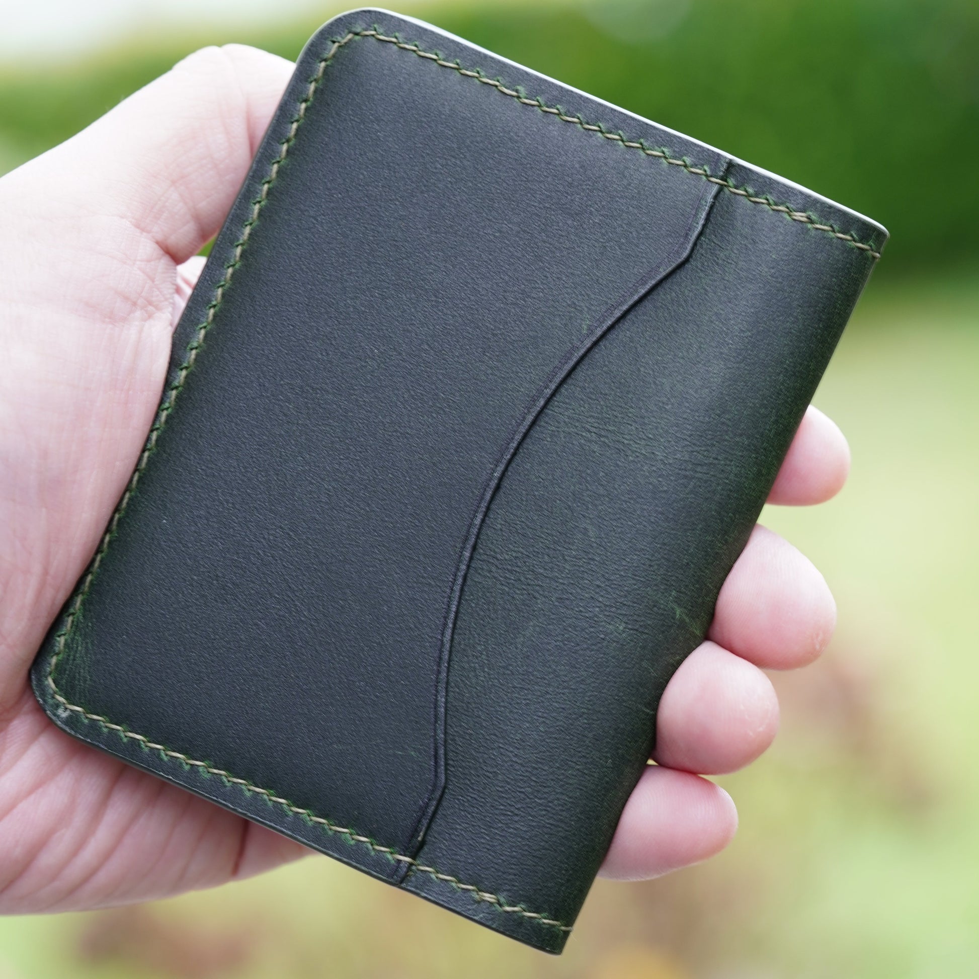 Hand holding a green leather card holder with green stitching and curved card slot.