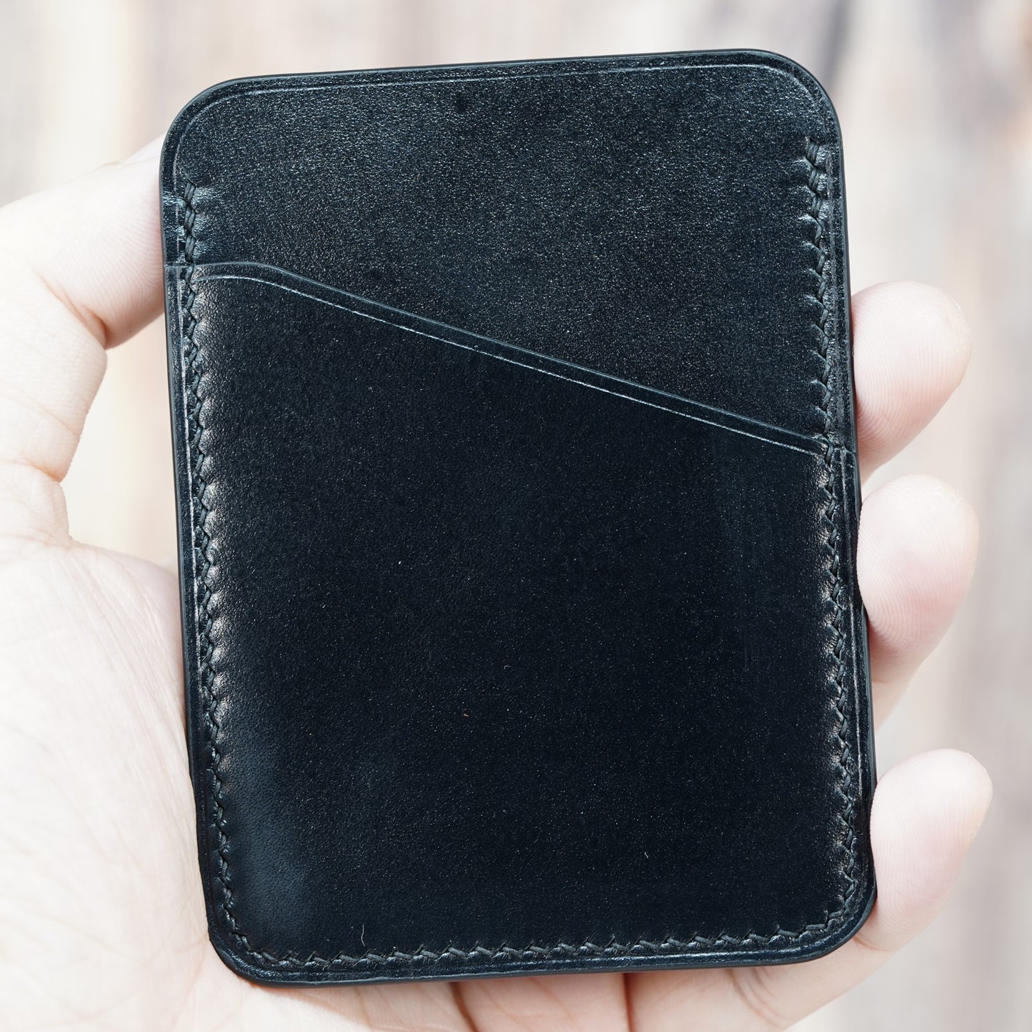 Hand holding a black minimalist leather card holder with visible stitching and a front card slot.