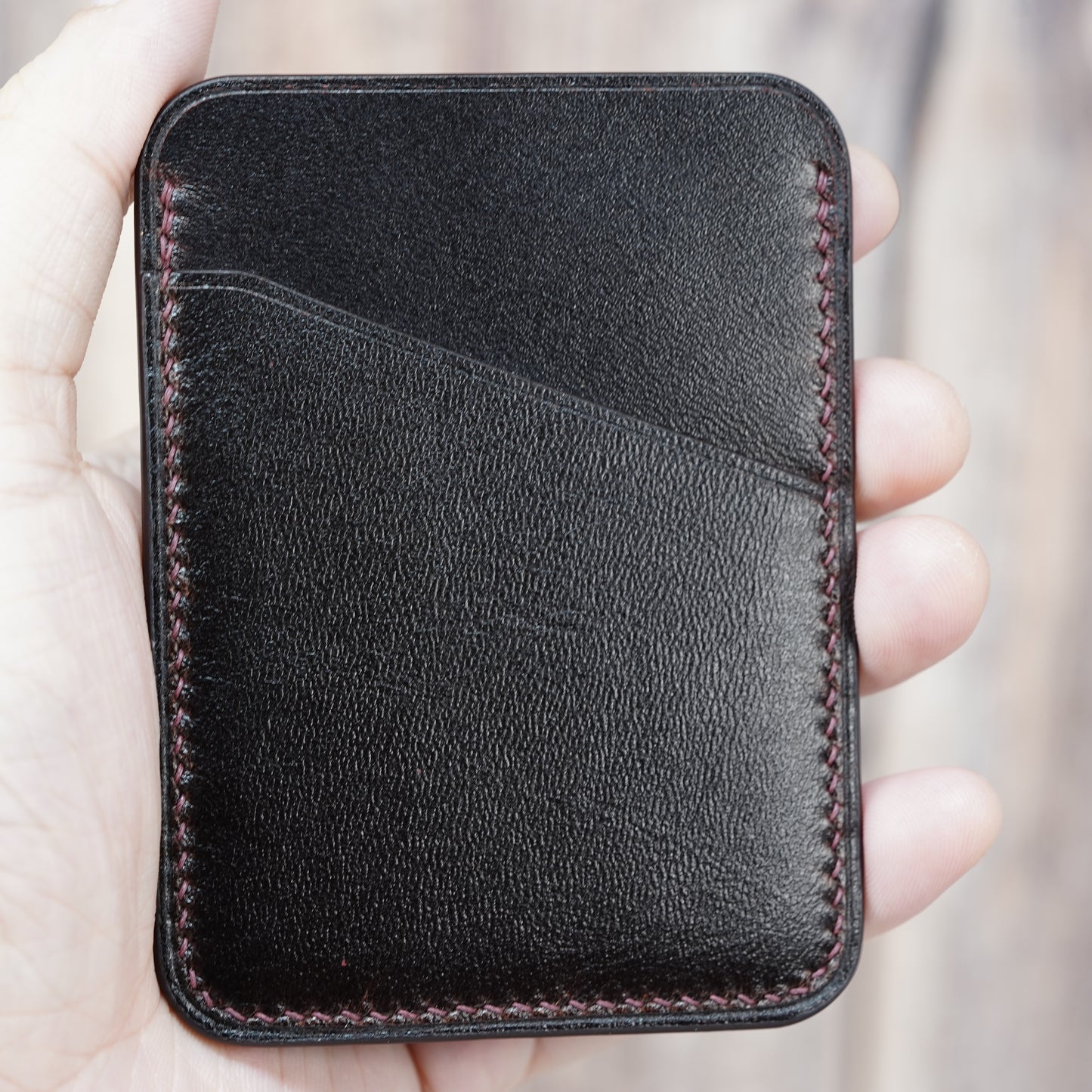 Dark brown minimalist leather card holder with red stitching, held in a hand.