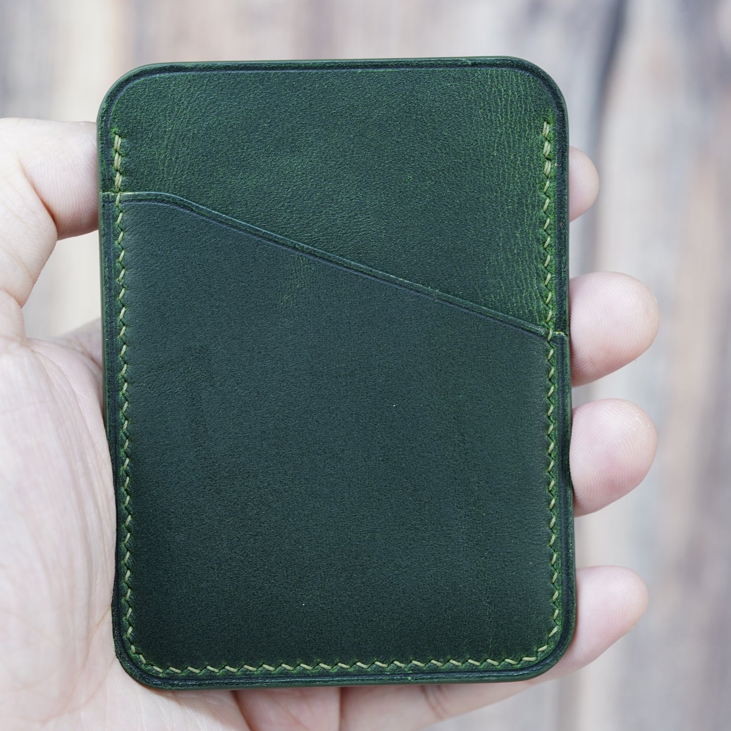 Hand holding a minimalist green leather card holder with light green stitching and two card slots.