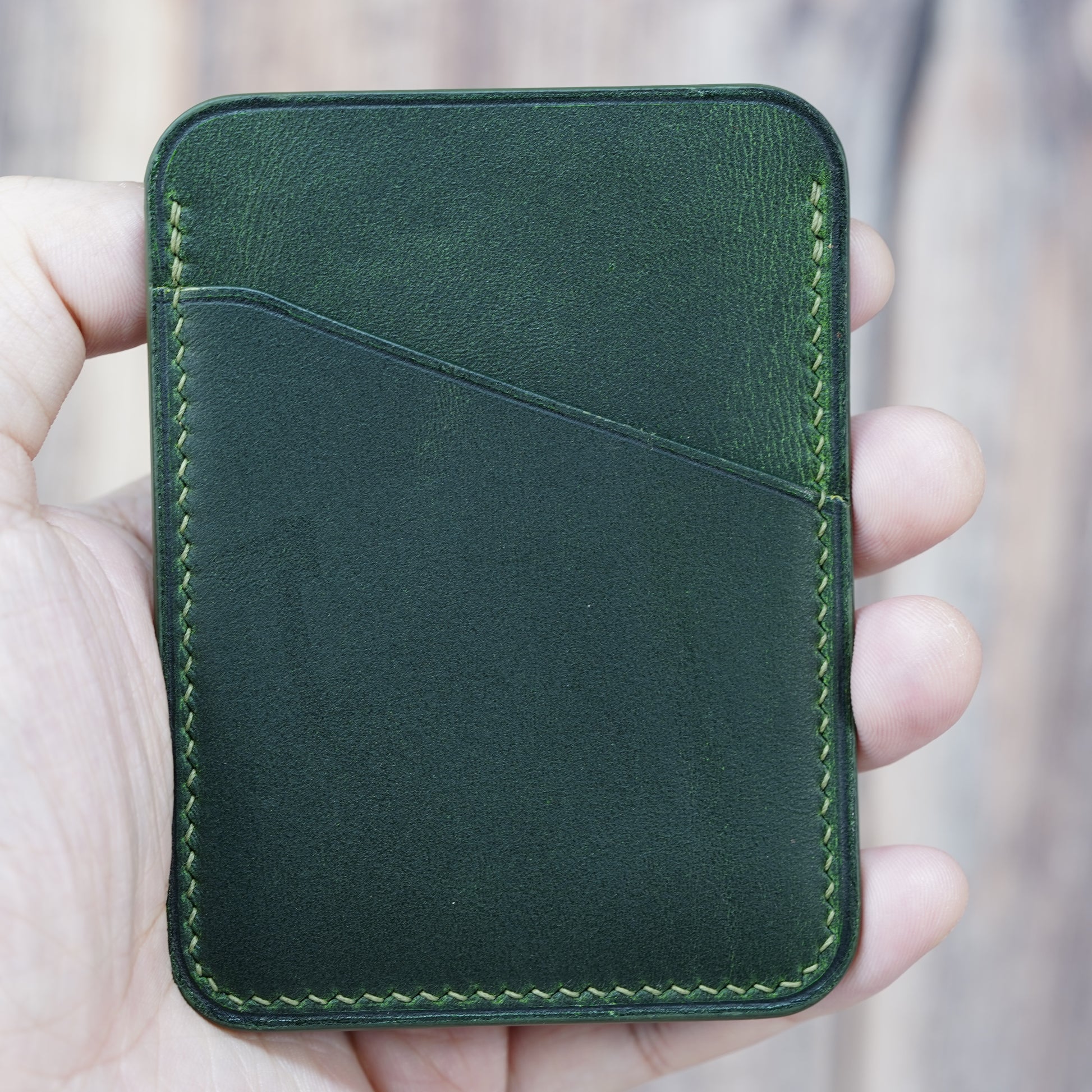 Hand holding a minimalist green leather card holder with light green stitching and two card slots.