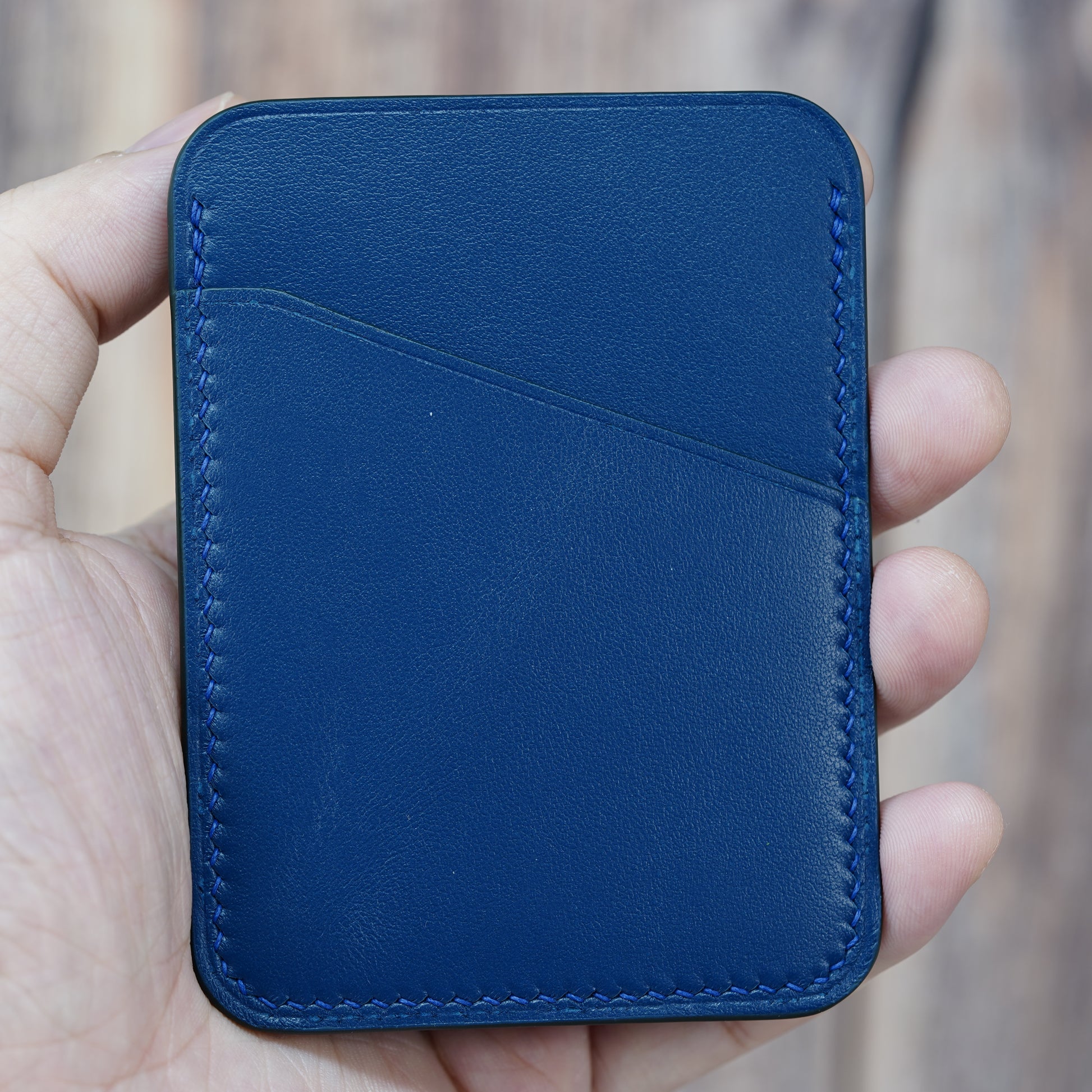 Hand holding a minimalist blue leather card holder with two pockets and blue stitching.