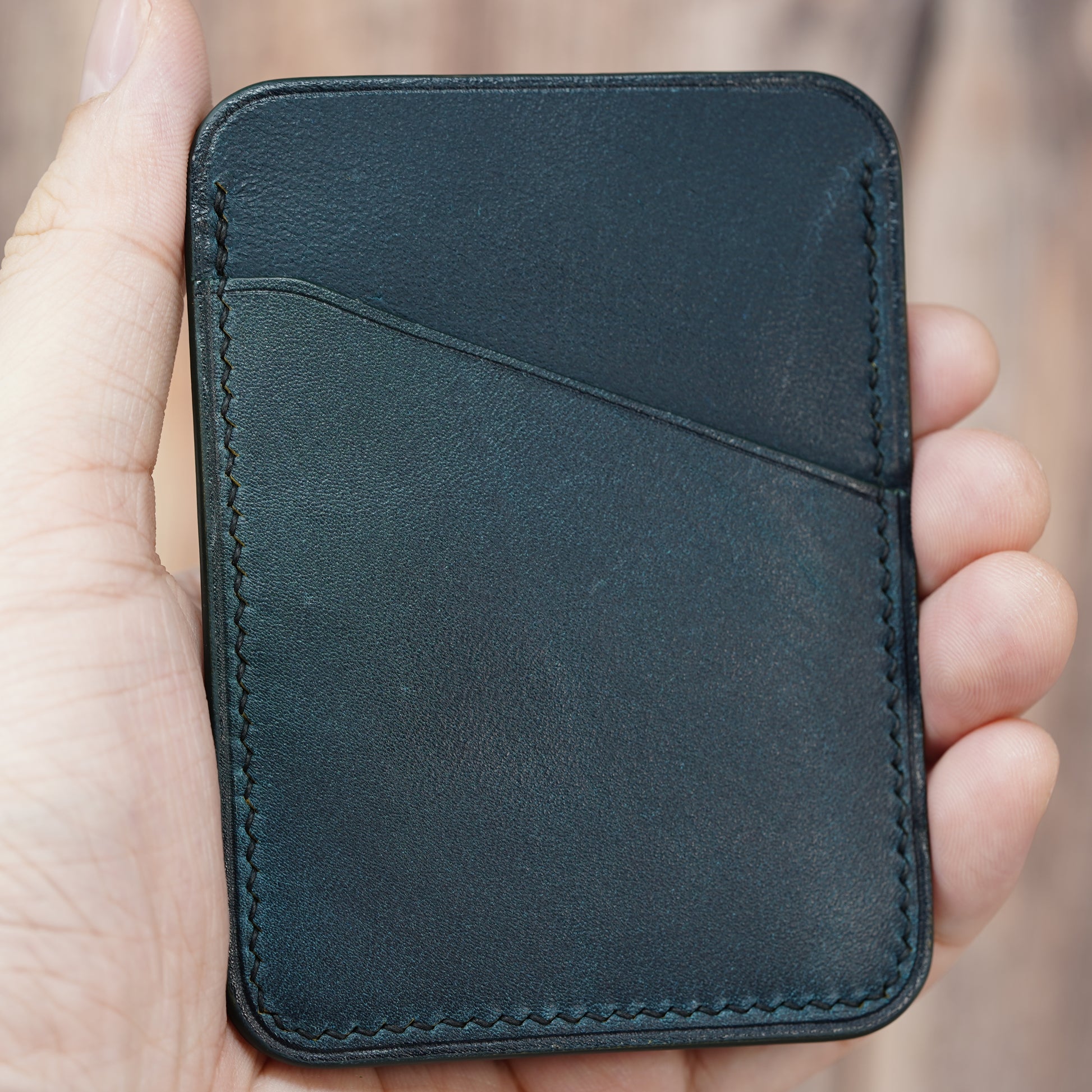 Hand holding a minimalist dark green leather card holder with diagonal pocket and visible stitching.