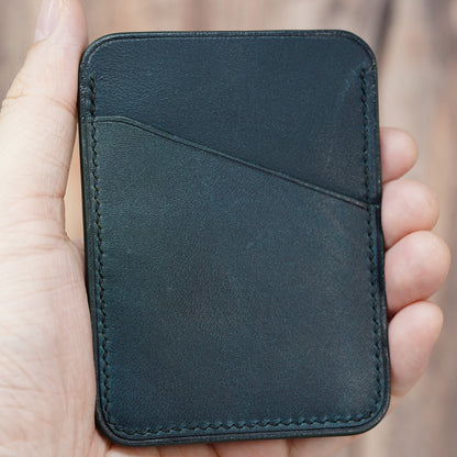 Hand holding a minimalist dark green leather card holder with diagonal pocket and visible stitching.
