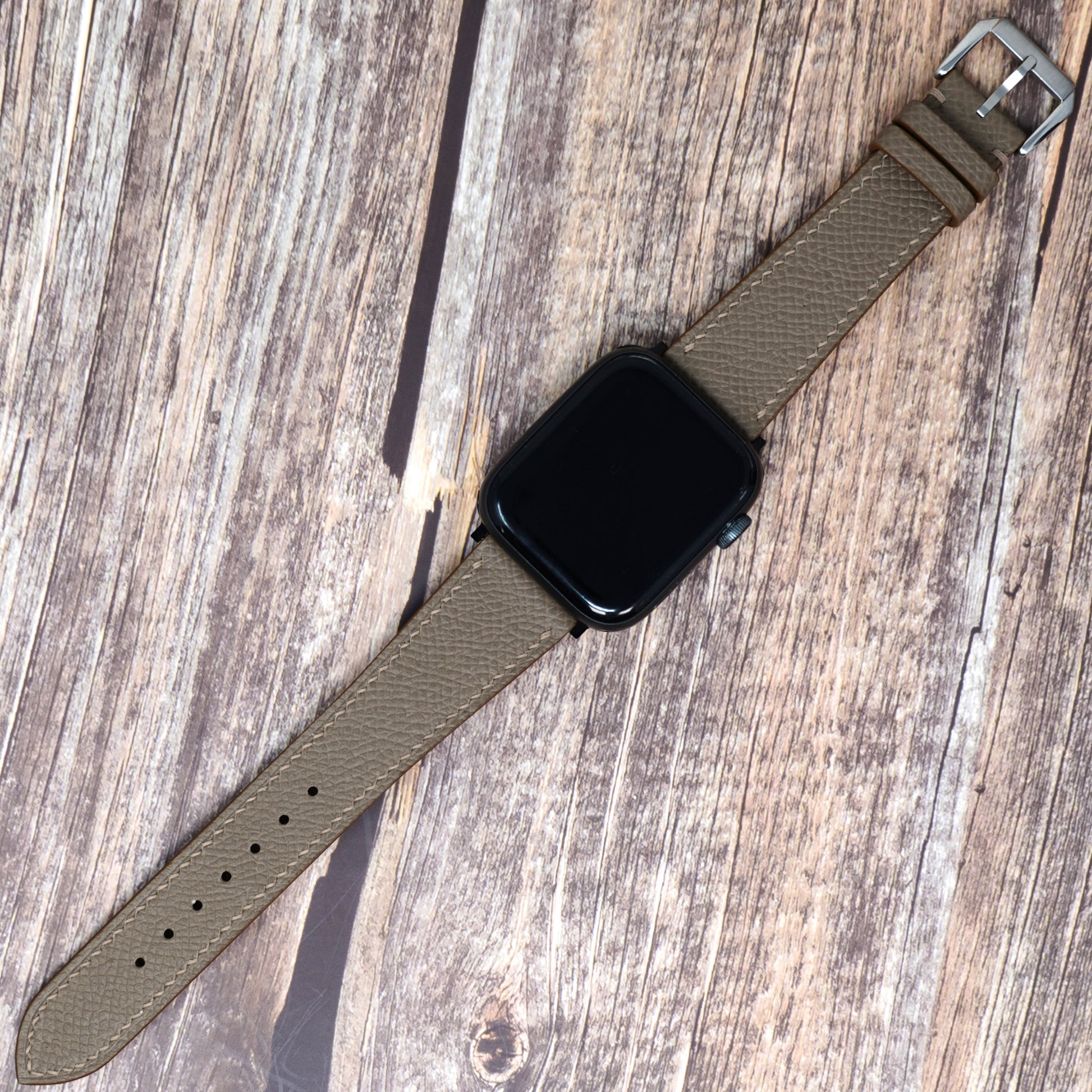 Taupe Epsom leather watch strap for Apple Watch, with a silver buckle, flat lay on wood.