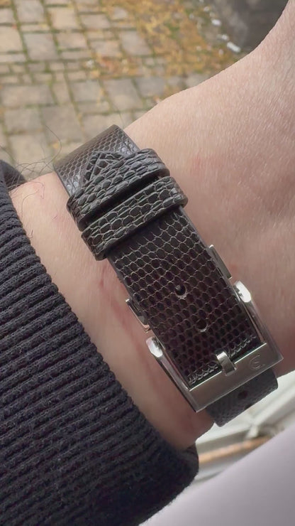 Dark brown lizard leather watch strap fitted to a steel dress watch, showing wrist fit, grain, stitching, buckle and underside.
