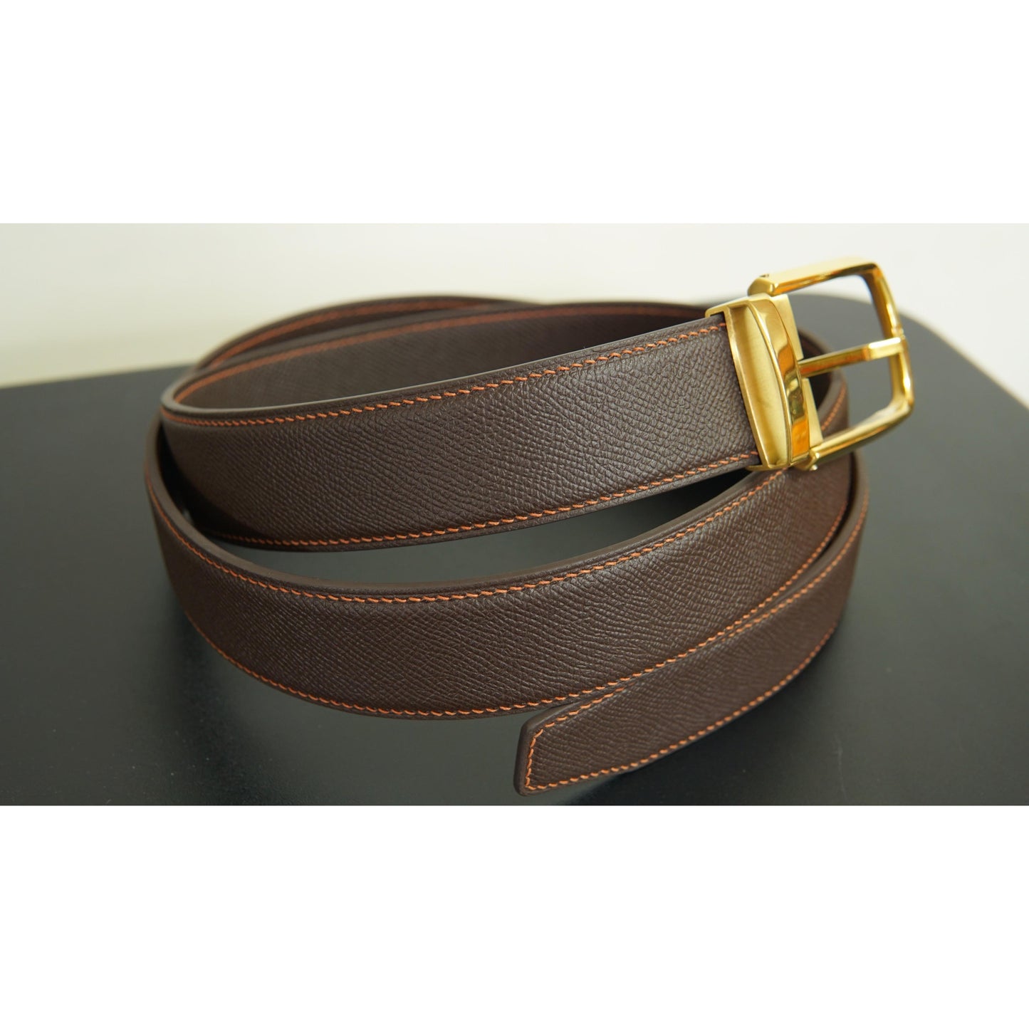 Brown leather belt with orange stitching and a gold buckle, coiled on a dark surface.