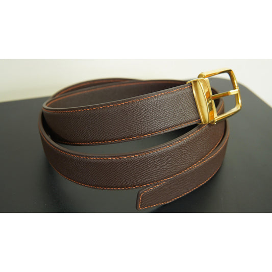 Brown leather belt with orange stitching and a gold buckle, coiled on a dark surface.